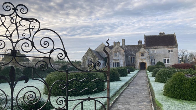 A stone mansion in winter; in front is a lawn with clipped topiary and a wrought iron gate